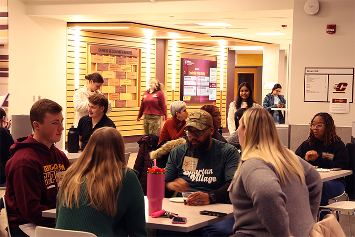Students gather in Grawn Atrium to meet their teams and practice their pitches for the Spark Challenge. In the foreground is a table with four students discussing their Impact Path prompt. Other tables have groups of students in conversation.