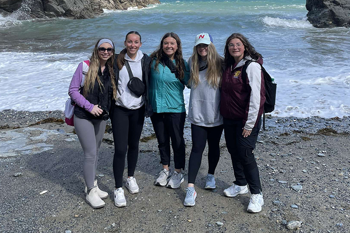 Five students stand on a beach in the United Kingdom. They are wearing jackets, sweatshirts, leggings and tennis shoes. The beach is rocky and waves roll in behind them.