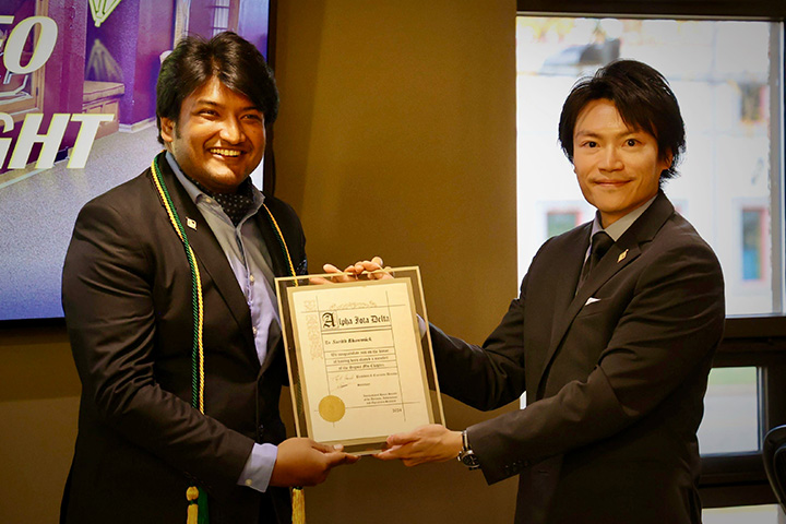 Surith Bhowmick receives an Alpha Iota Delta certificate from Edward Chou. Both are dressed in jackets and Bhowmick wears green and cold cords and tassels.