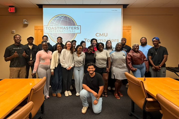 CMU Toastmasters members pose together in a conference room in front of a projected Toastmasters International logo.