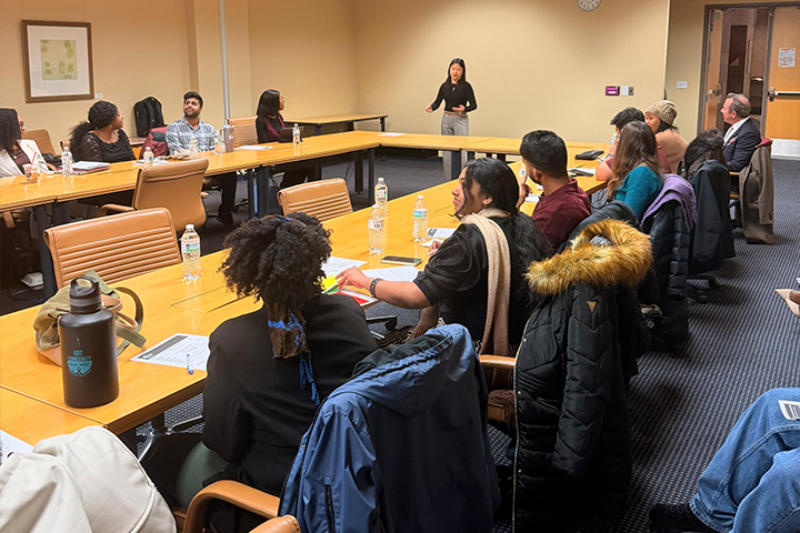 A student speaks at the front of a classroom while peers sit around a conference table during a CMU Toastmasters meeting.