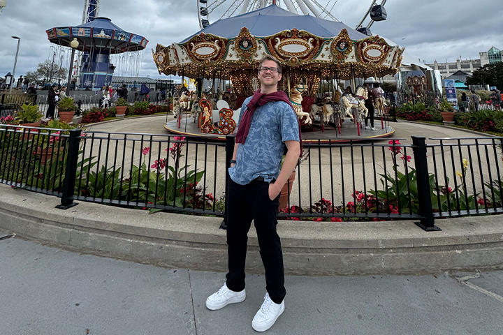 Trevor Carnovsky wears a blue t-shirt with black pants. a maroon long sleeve shirt tied around his neck, and white sneakers and poses with his hands in his pockets. Behind him is a carousel and ferris wheel.