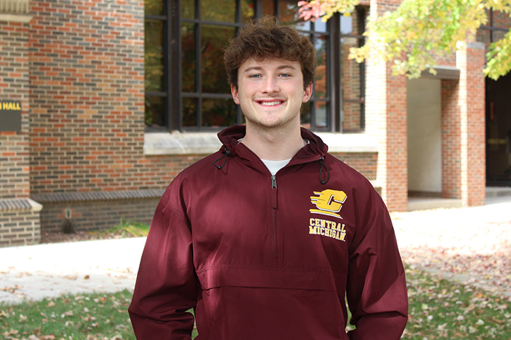 Tyler Harvill smiles outside Grawn Hall at Central Michigan University, wearing a maroon CMU jacket with the university's gold logo.