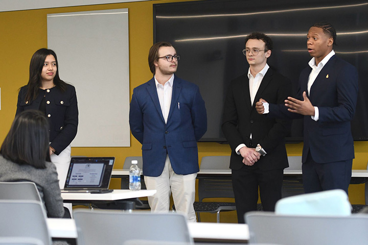 Derek Ballas, Blake Jimenez, Carolina Hernandez Ruiz and Mark-Allen Gay stand in front of ventures seeking funding at the Wayne State VC competition. They are pitching to the ventures and gesturing as they speak.