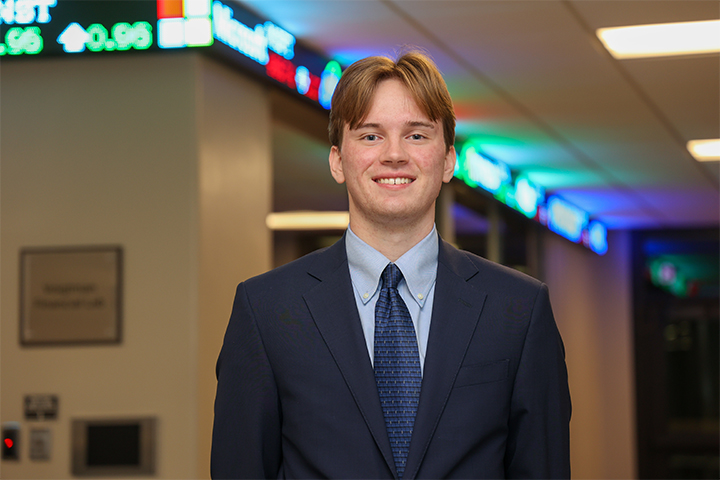 Wyatt Ross, a Central Michigan University finance student, stands smiling in a suit outside the Voigtman financial lab with digital stock displays in the background.