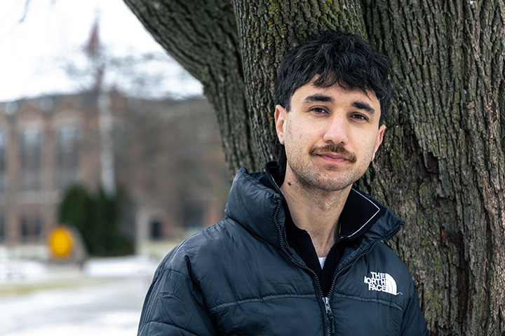 Zane Iqbal stands against a tree on the Warriner mall during the winter. He's wearing a black North Face jacket over a black collared shirt. He has dark hair and a mustache.