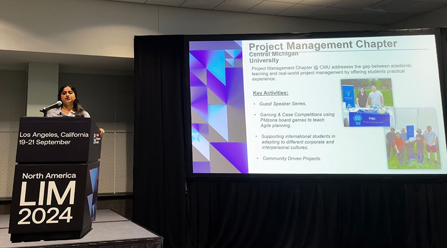 Naina Rajan stands at a tall black podium with North America LIM 2024 on the front in large white letters. Behind her is a projector screen showing a slide on the Project Management Chapter.
