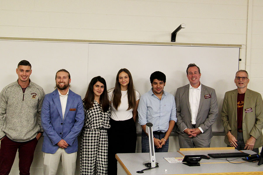 Clay Kosnik, Casey Croad, Aimen Khan, Sarah Firchau, Priyanshu Sharma, Pres. Neil MacKinnon and Jeff Hoyle stand in a line across the front of a dry erase board in a CBA classroom. They are looking at the camera and smiling.