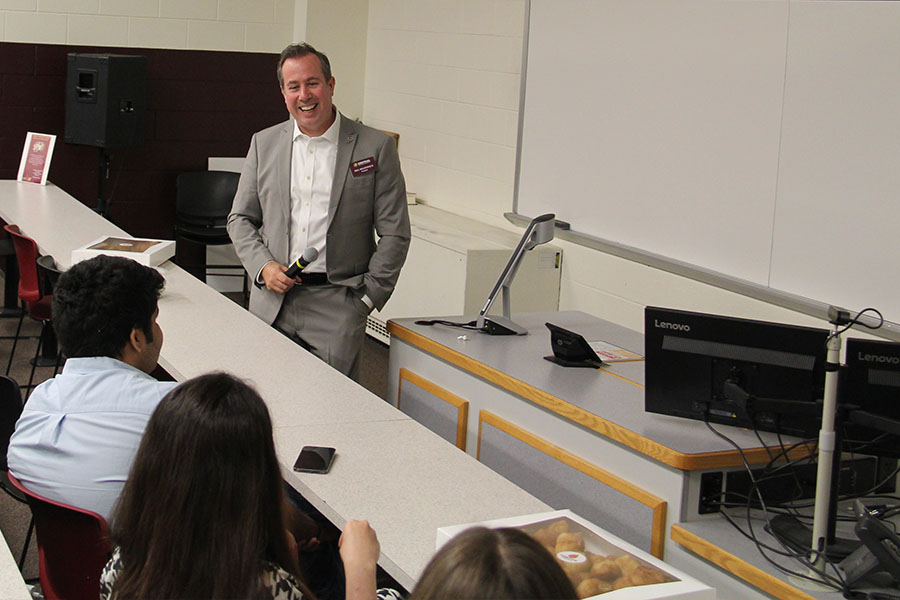 President MacKinnon holds a microphone as he laughs. He's wearing a gray suite with a white shirt and his maroon nametag. In front of him sitting at desks are students attending the AMA launch event. There is a box of donuts on the desk.