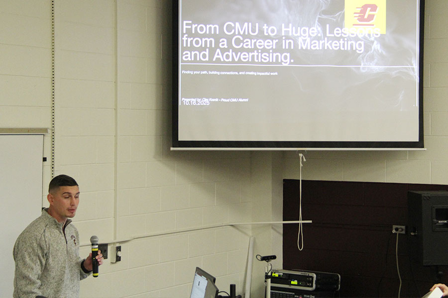 Clay Kostnik wears a gray long sleeve pullover with a half zipper and a CMU logo. He stands in front of a pull down screen with a PPT slide that reads