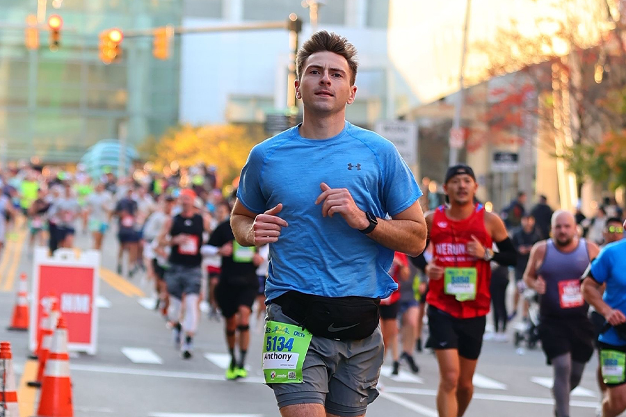 Anthony Niemiec wears a blue shirt and gray shorts with a black waist bag. He has a race bib on his leg. Behind him are many other runners on a city street in Detroit during the Detroit Marathon.