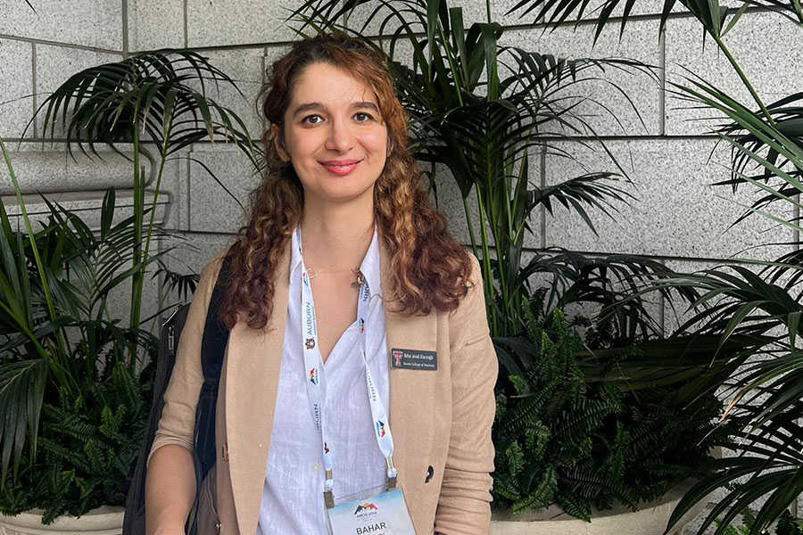 Bahar Javadi Khasraghi stands in stone hallway in front of palms and ferns. She has a tan jacket with a name tag, a conference badge, and a purse over her shoulder. She's smiling at the camera. 