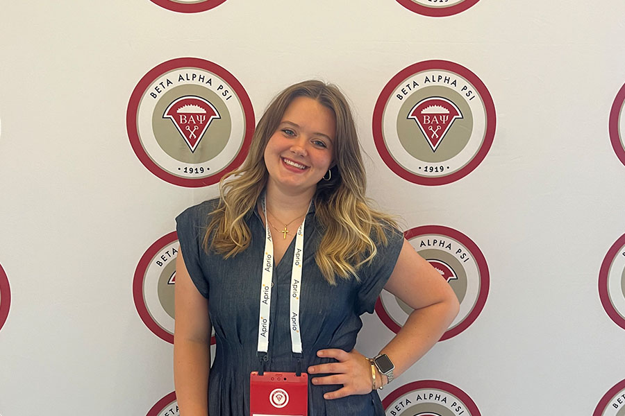 Bailey Graham stands in front of a Beta Alpha Psi step-and-repeat logo banner. She has long light brown hair with blond ombre. She's wearing gold loop earrings, gold necklaces, gold bracelets, and a watch. She wears a light blue cap-sleeve dress.