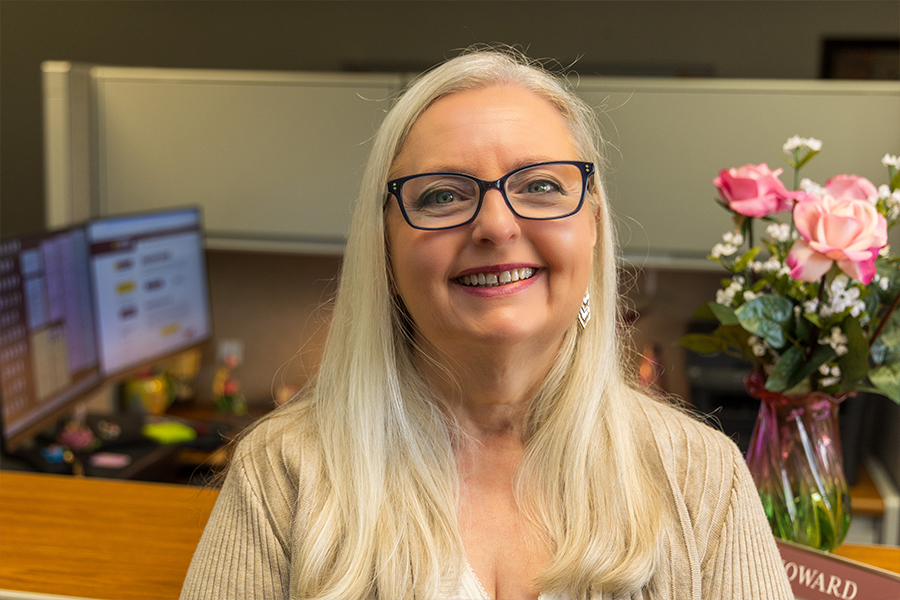 Cindy Howards stands in front of her desk with a vase of pink roses behind her. She has long white and gray hair, black glasses and silver earrings. She's wearing a tan cardigan over a white tank top.