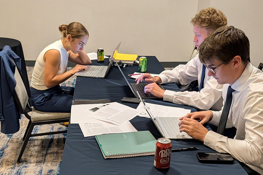 Savana Stewart, Austin Benchley and Zach Parker sit at a long table covered with notebooks, paper and soda cans. They peer intently at their laptops. They wear business professional cloths, but their jackets have been removed.