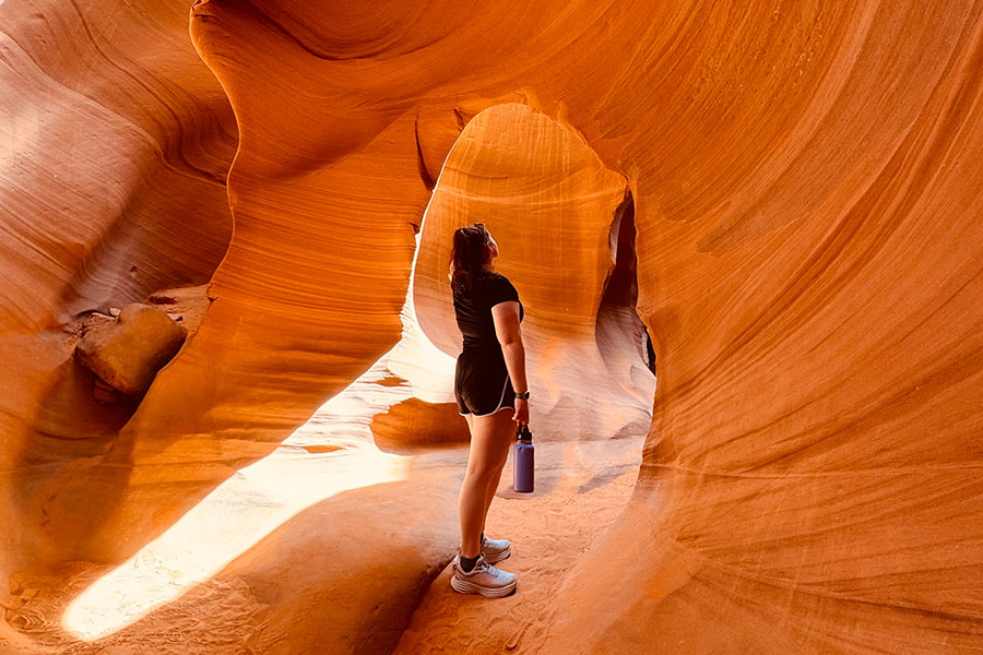 Corinne Sleeter faces away from the camera in black shorts and t-shirt, holds a water bottle and looks at sandstone formations in the desert.
