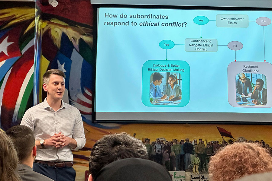 Adam Effinger stands in front of a classroom. He's wearing blue slacks and a white button down shirt. Next to him is a pull down screen with an image of a flow chart on ethical conflict.