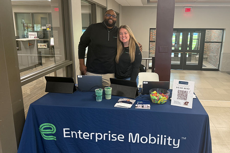 Daniel King and Julia Annarino wear black long-sleeve zip up shirts and stand close with their arms around each other as they post in front of the recruiting table next to Business Student Services. The table has a blue Enterprise Mobility tablecloth.