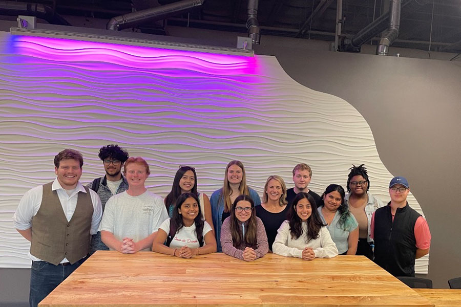 Twelve students and their advisor Julie Messing stand at a natural wood table. The students are of mixed genders and races. Behind them is a white textured wall with ripples and a neon purple light.