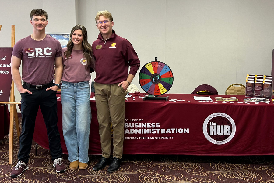 HUB Ambassador Ethan Rauschert wears a CMU branded maroon pullover and khakis and stands with two other students in front of a table with a maroon CBA table cloth and giveaways.