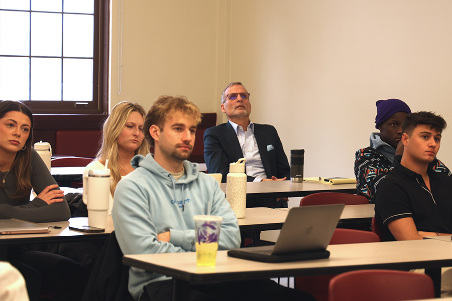 Jeff Hoyle sits in the third row of a classroom. He is waring a dark suit jacket with a light blue pocket square. Five students fill the seats in front of him. They are all looking forward intently at something happening in the front of the class.
