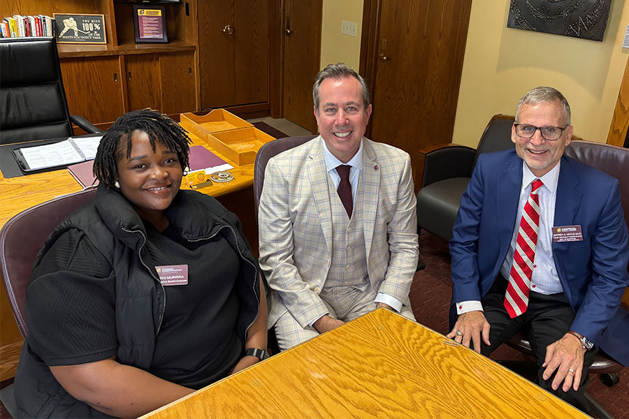 Farirai Murwira has short black dreads, silver earrings, and wears a black short-sleeve shirt and black vest and sits next to President MacKinnon and faculty member Jeff Hoyle. They sit at a large wooden table in MacKinnon's office.