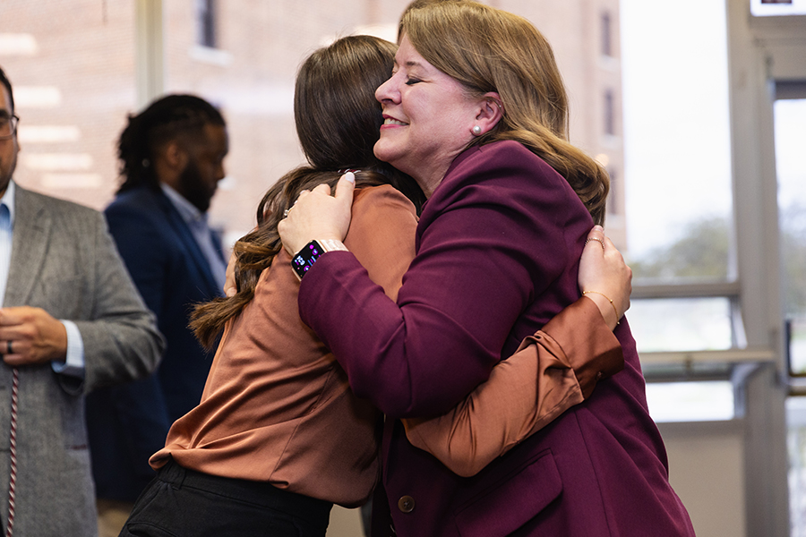 Dr. Amy McGinnis in a maroon blazer hugs first-gen student Madison Zellar in a tan long-sleeve shirt at the first-gen cording ceremony. A man in a gray blazer holds a maroon and gold cord in the background.