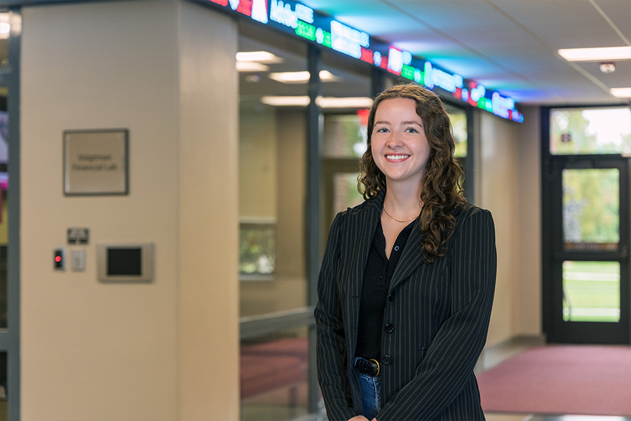 Holly Ireton stands outside the Bloomberg Lab in Grawn Hall. The news scroller is brightly lit above her. She's wearing a gold necklace, a black and gray pinstriped blazer over a black shirt. She has long brown curly hair, blue eyes and a big smile.