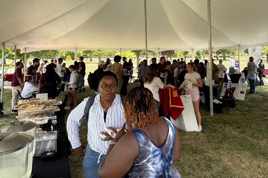 Students gather under a large white outdoor tent. There are tall tables with information from various student organizations.  On the left is a table with drinks and snacks. There is a large crowd of students looking at the different tables. 