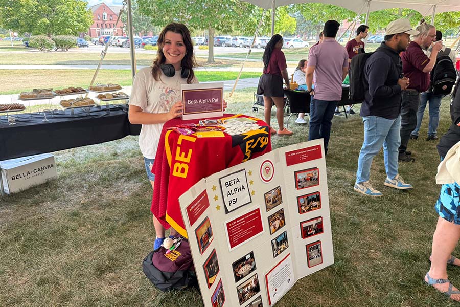A Beta Alpha Psi representative stands at the BAP table and smiles. She's wearing shorts and a white tshirt. There is a BAP flag draped over the table. In front of the table is a posterboard with photos of the group. Students stand in the background.