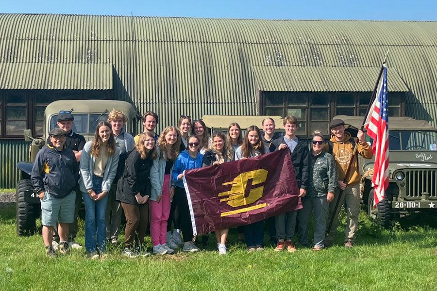 Jerry DiMaria (far left) stands with a group of students and faculty member Michelle Bartosek in front of a military building and three military jeeps. The groups is dressed for cool weather. They hold a CMU flag and an American flag.