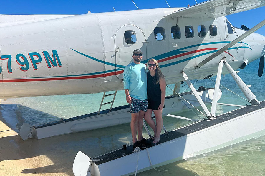 Julia and her husband stand in beach clothes on a ramp leading to a boat plane floating in water on a sunny day..