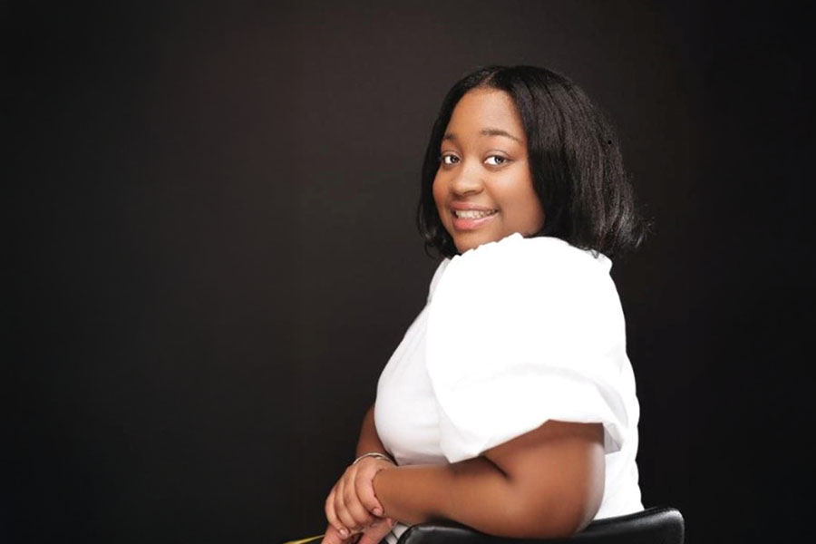 Kamaria Milton wears a white shirt and sits in a black leather chair. She has her arms clasped to the side and she's posed looking back over her shoulder with a big smile. The background is dark.