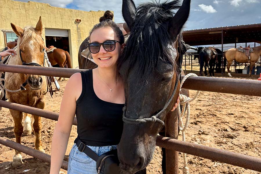 Kayli Kalish wears a black tank-top and jeans and stands next to a dark brown horse.