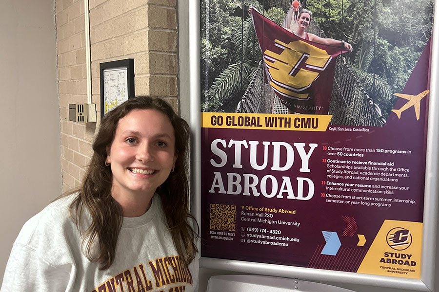 Kayli Kaylish wears an off-white CMU sweatshirt. She has long brown hair and smiles as she stands next to a large Study Abroad poster in Grawn Hall. The poster features a photo of Kayli holding an Action C flag on a rope bridge in a Costa Rican jungle.