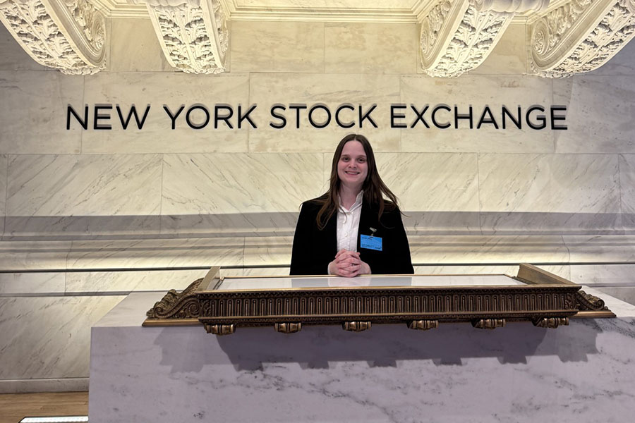 Kylea James smiles and stands under the NYSE bell wearing a black business suit with a white button down shirt.