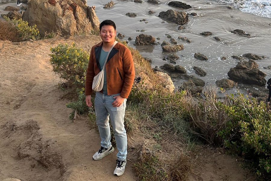 Liu Zhanning wears light blue jeans, white tennis shoes with black laces, a blue shirt, and a tan jacket. He has a tan cross-body bag. He's standing at a beach with sand and scrubby plants. There are rocks behind him.