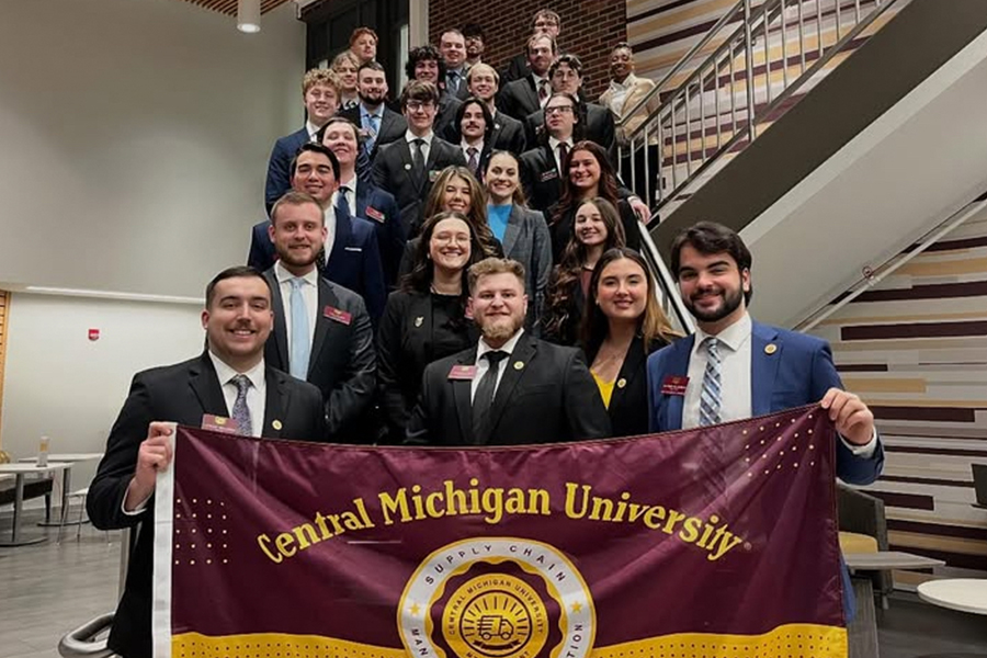 A group of nearly 30 students stand on the stairway in the Grawn Atrium. They are dressed in business professional clothing. The bottom two students hold a CMU Supply Chain flag that is maroon and gold.