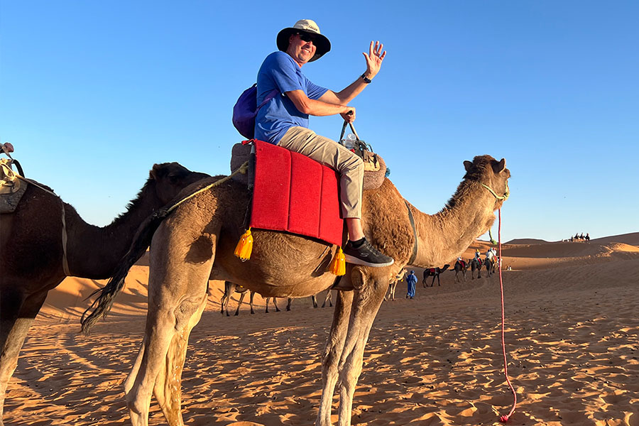 Lyndell Patrick wears a blue tshirt and a pair of khakis as he sits atop a camel in the desert.