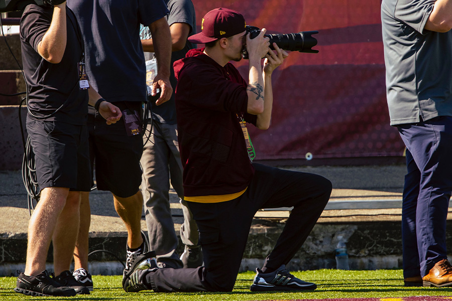 Mac Mayhew kneels on the sidelines of a CMU football game. He has his eye to a large SLR camera. He has his maroon CMU baseball cap on backward and wears a maroon shirt over black pants with athletic shoes. A press pass hangs around his neck.