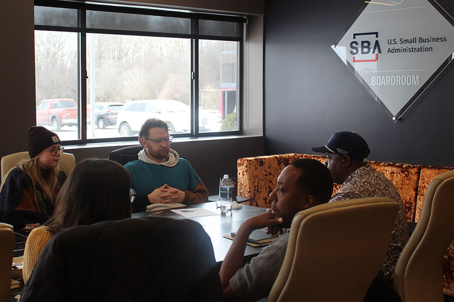 Students in sweatshirts and sweaters and mentors sit around a table in a Small Business Administration boardroom. A window is behind them showing a parking lot.