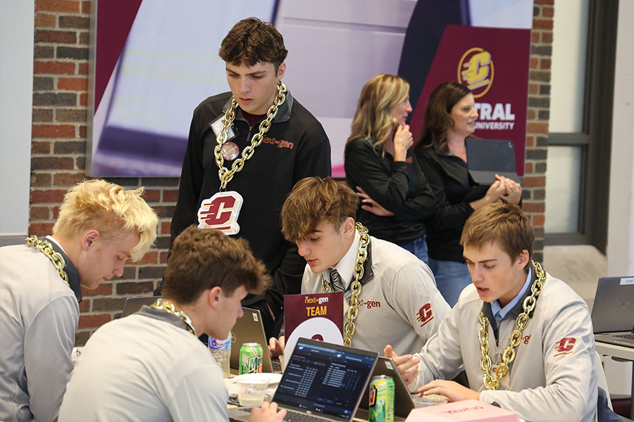 Four students in tan long-sleeve Next-Gen branded shirts with Action C logos wear large gold plastic chains with Action C logos. They sit around a table looking at laptops. A mentor wears a matching black shirt, gold chain and stands next to them.