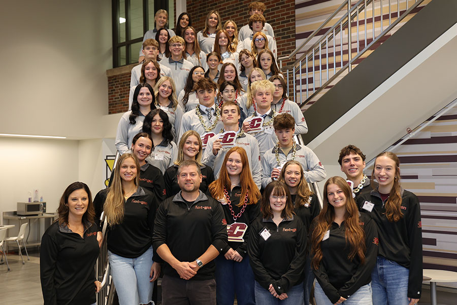 High school students stand with their mentor and BIS faculty members on the Grawn Atrium stairs. They are wearing matching NextGen and CMU branded longs-sleeve shirts. Some wear the large chains with Action C logos.