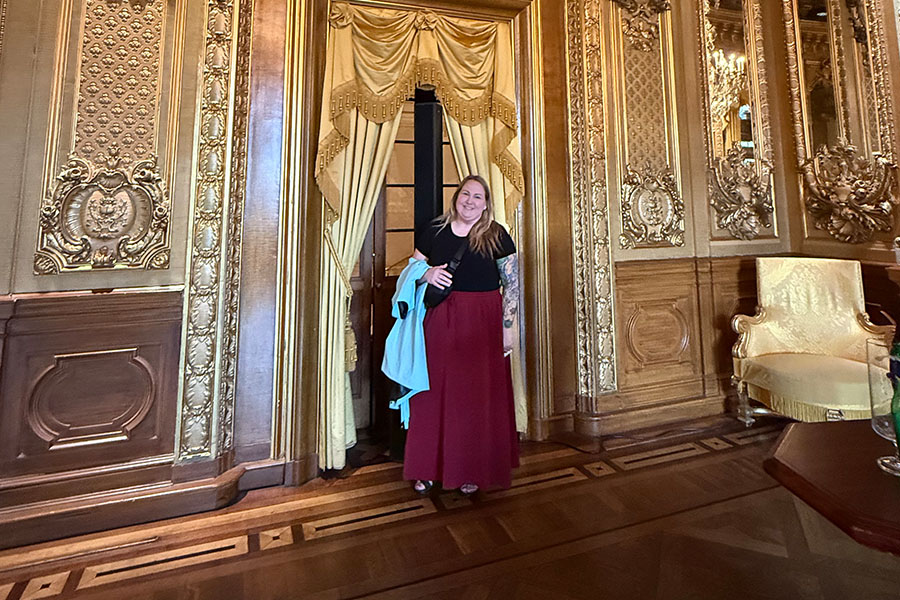 Nicole Erckman stands in a Swedish opera house with ornate gold embellishment sand dark heavy wood. Behind her is a gold satin curtain with gold ropes. She's wearing a black short-sleeve shirt and maroon wide-legged pants with heels.