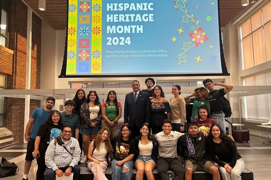 A large group of Prospanice group members pose with a guest speaker under a colorful Hispanic Heritage Month 2024 display banner in the Grawn Hall Atrium.