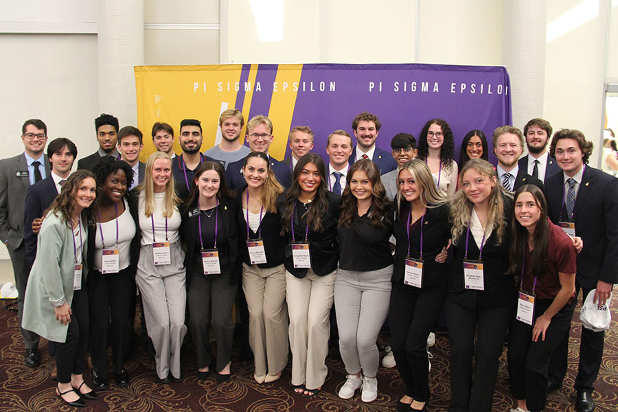 A large group of students in business clothes wearing nametags on lanyards pose in front of a yellow and purple photo backdrop in the UC rotunda.