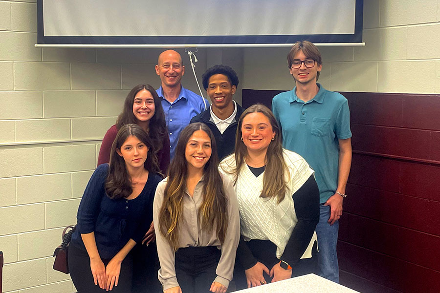 Four women students and four men students stand with their faculty advisor in a classroom during the inaugural meeting of Rho Epsilon Iota. They are wearing business casual clothing and posing as a group in two rows with the advisor directly behind them.