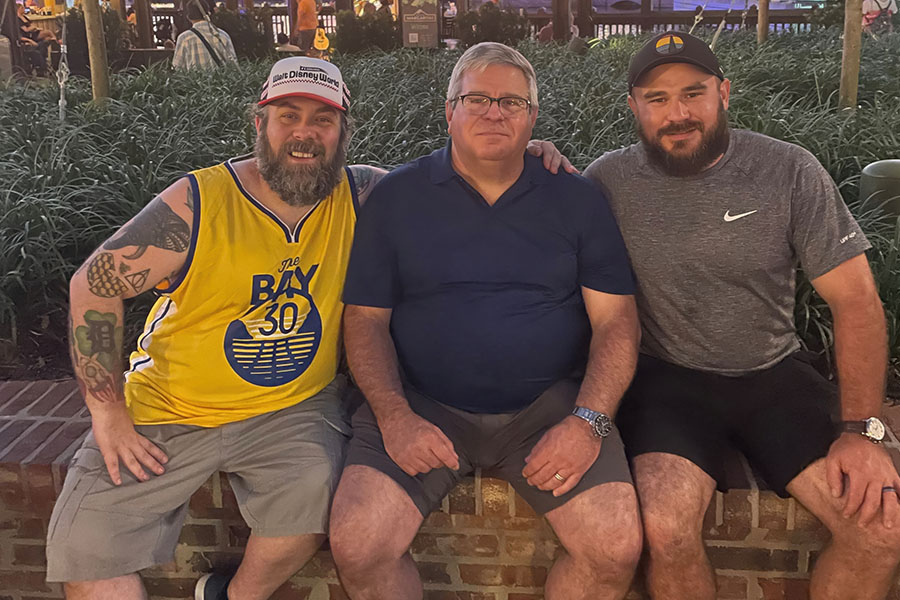 Rick Wright, his father and his brother wear t-shirts and shorts and sit on a short brick wall with decorative grass behind them.