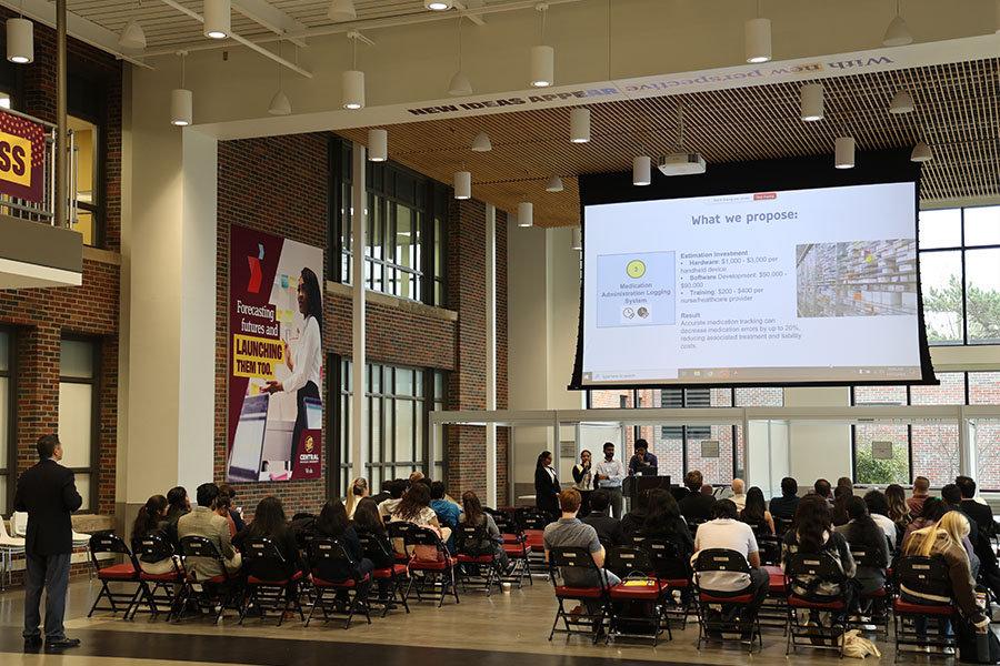 A group of four students stands in front of a seated audience in the Grawn Hall atrium. They have a PowerPoint slide displayed on a large screen above them.