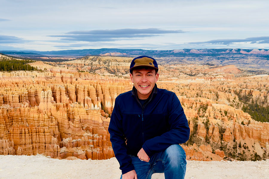 Ryan Murphy crouches in front of sandstone hoodoos at Zion National Park. He is wearing jeans, a blue jacket and a blue and tan hat. He's smiling and has an arm on his knee.